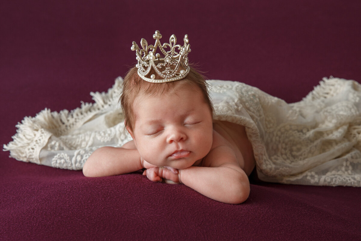 Portrait of a beautiful baby girl wearing a princess crown and laying on a purple blanket with a white cover