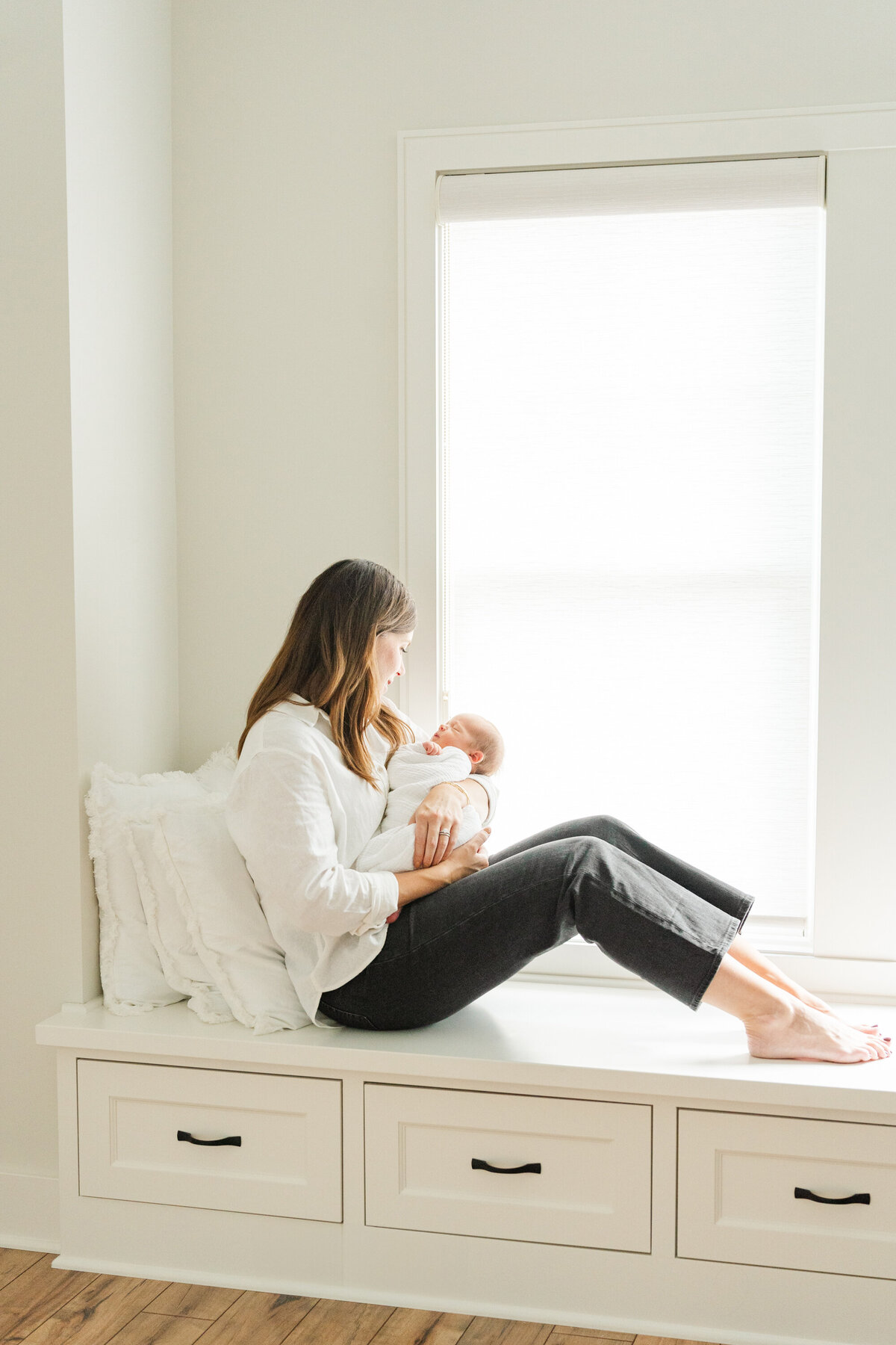 A mother sits in a cozy window nook, lovingly holding her newborn baby in her arms