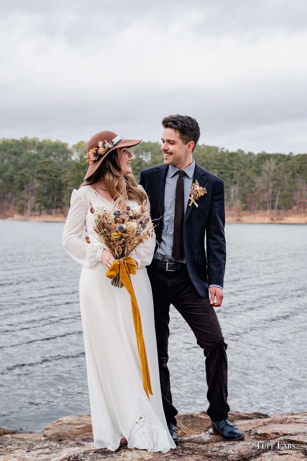 A bride and groom look at one another and smile as she holds her dried flower bouquet and water is seen in the background.