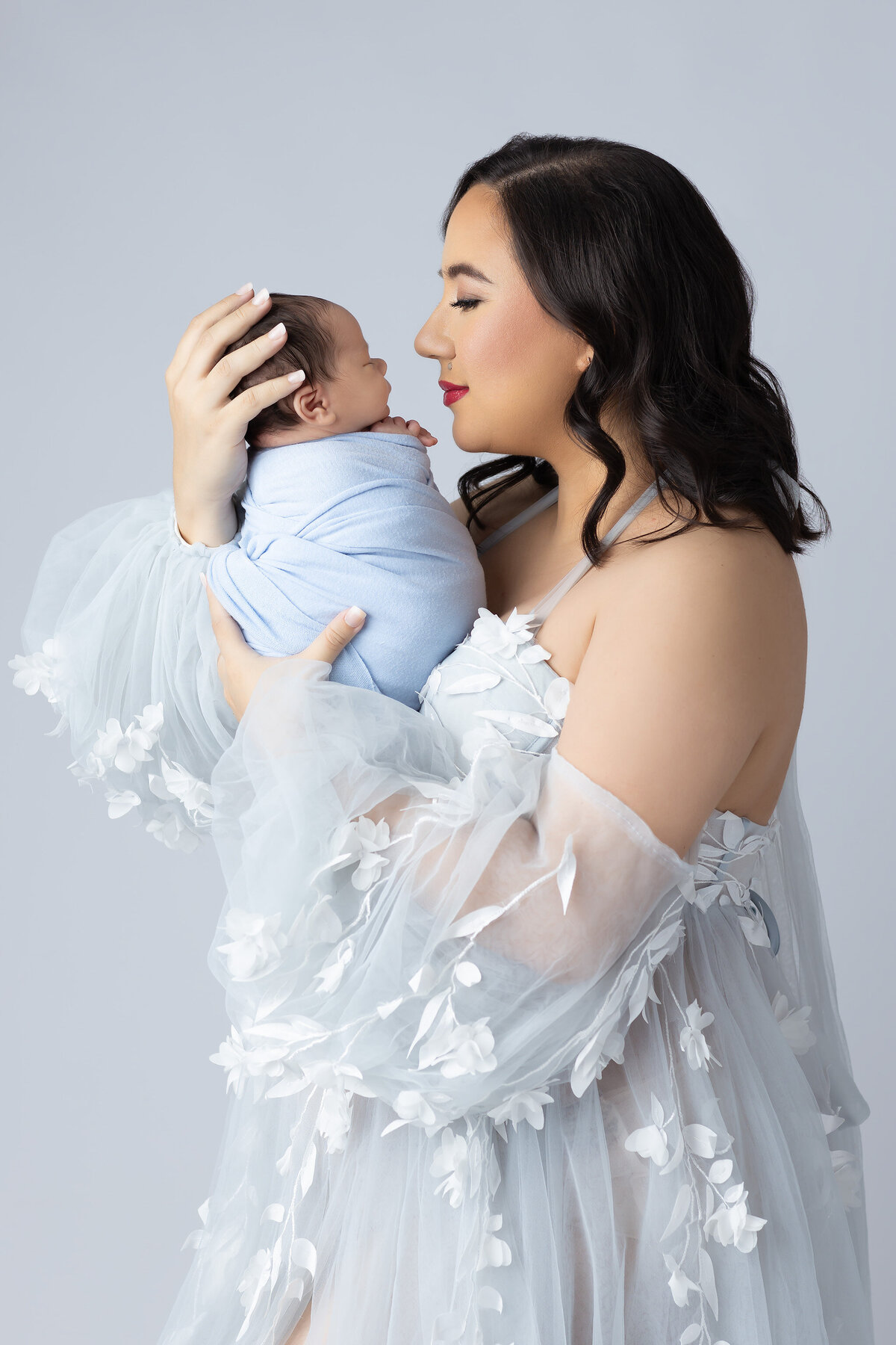 Elegant mom in a sheer embroidered gown holding her newborn wrapped in blue.