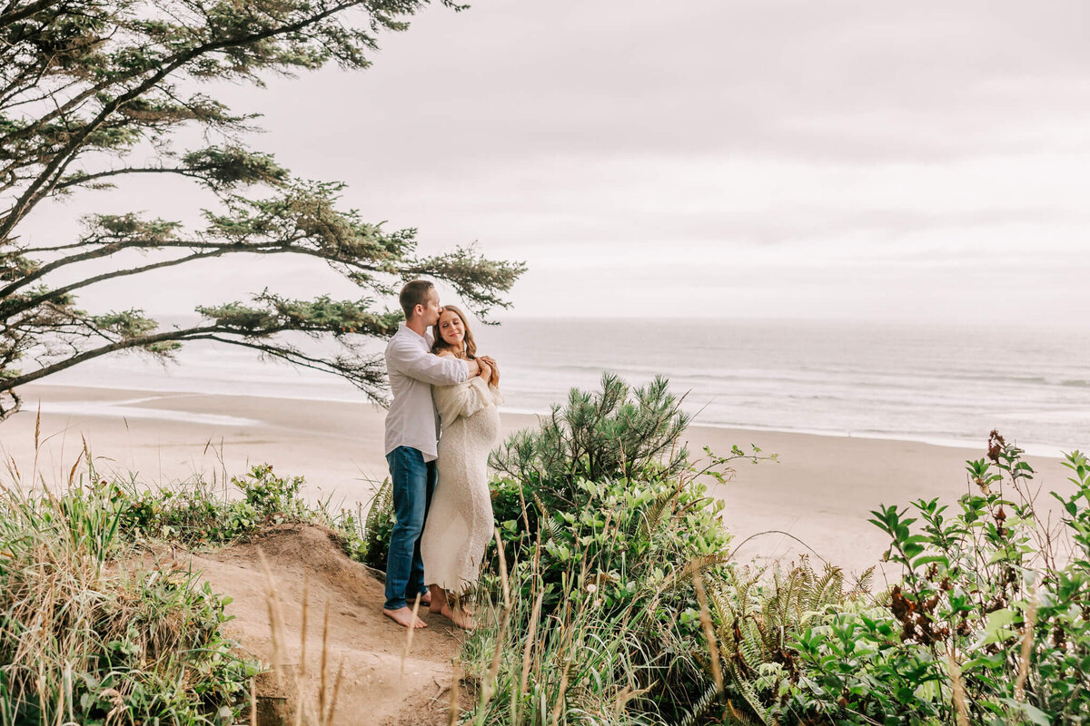 Portrait of a woman and her husband hugging on on overlook at the oregon coast. She is pregnant and wearing a cream dress.