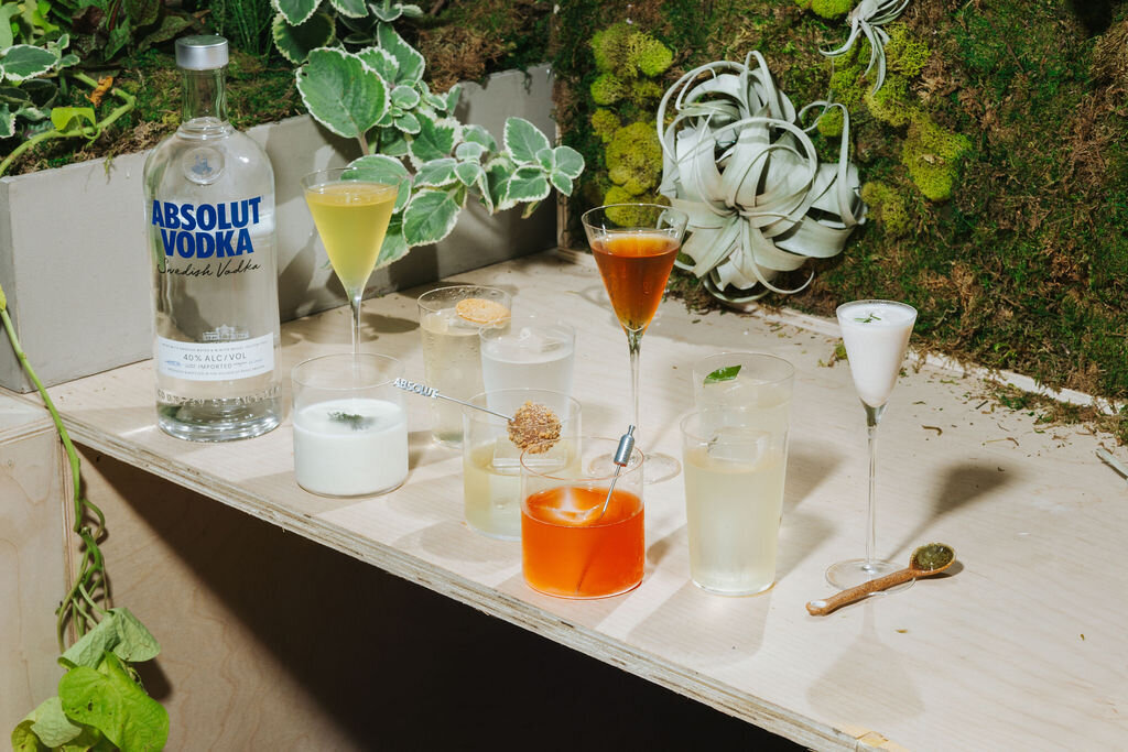 A bar counter styled with Absolut Vodka bottles and colorful cocktails surrounded by greenery.