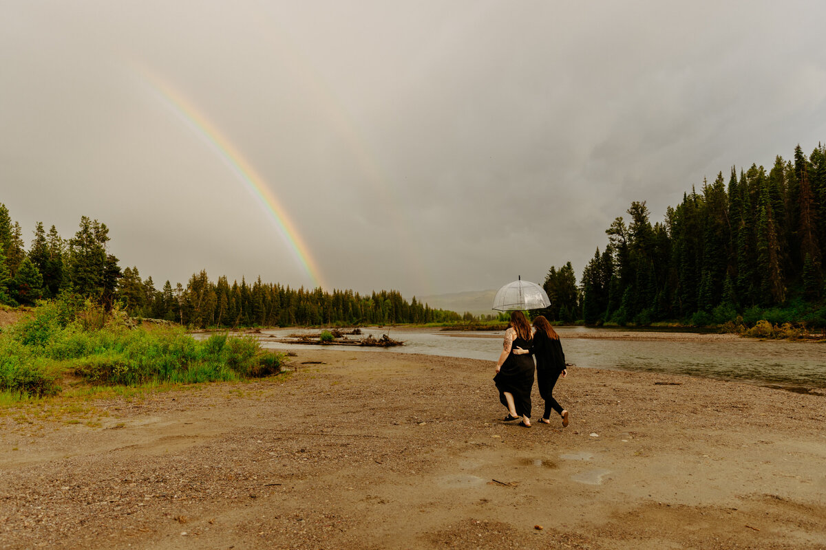 a lesbian couple walking into the distance under a double rainbow cuddled under an umbrella