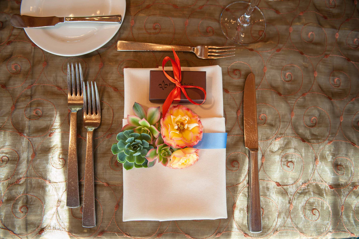 place setting with flower napkin treatment of succulent, orange dahlia, and blue ribbon wrap