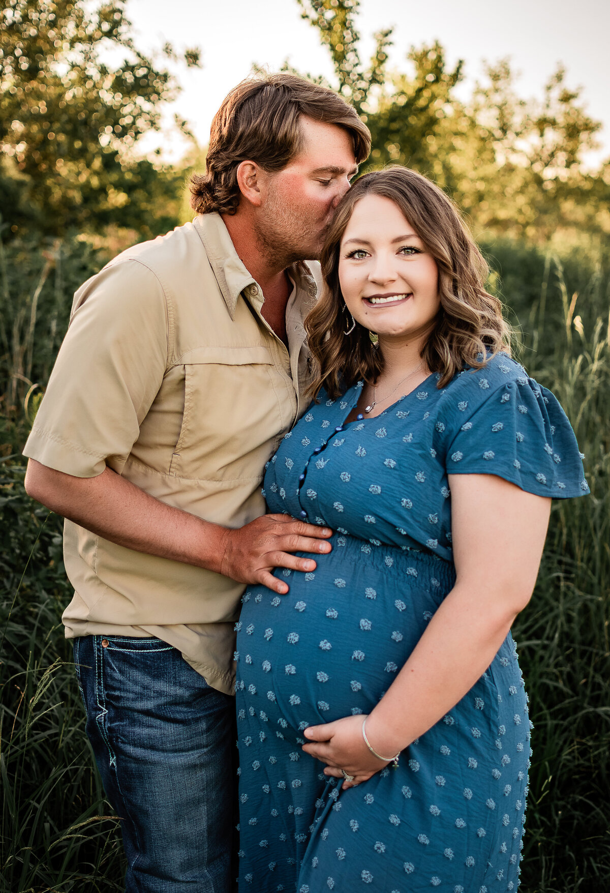 An mother to be smiles at the camera as her husband kisses her cheek and cradles her baby bump.
