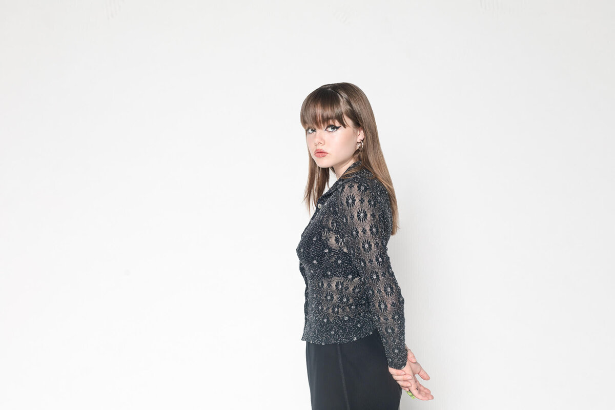 A woman with long hair stands against a plain white background. She is wearing a black lace top and black pants, looking to the side with her hands behind her back.