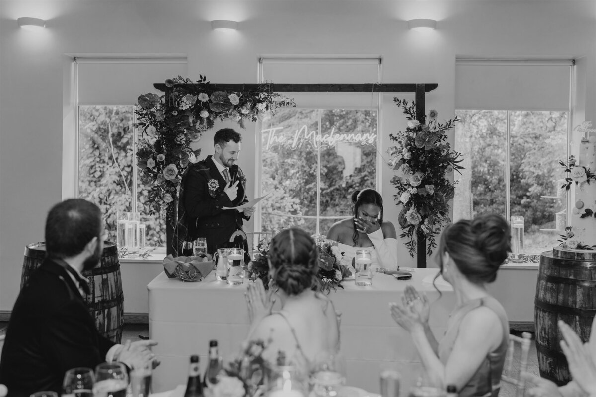A groom stands in front of a large floral display and holds a piece of paper. He smiles and deliver a speech. Guests clap and watch him speak.
