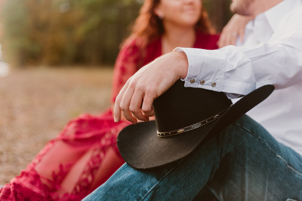 Rustic engagement photo of Cynthia and Leon in Humble, Texas. The couple embraces, with Leon holding a cowboy hat, and Cynthia in a flowing red dress. Captured by ShutterUp Photography, specializing in engagement and couples sessions.
