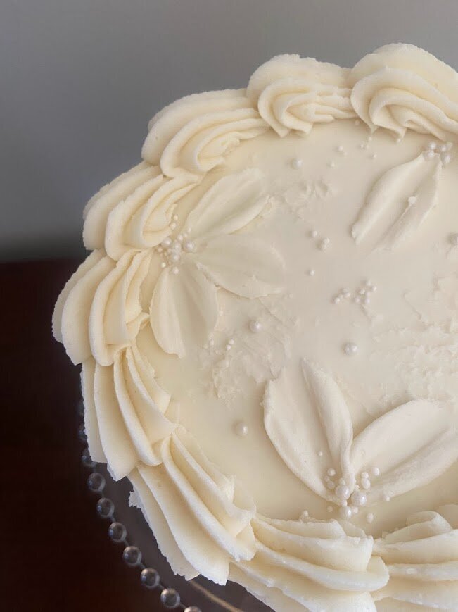 Photo shows the top of a white cake, with a shell piped border, and pallet knife florals with pearl sprinkles.