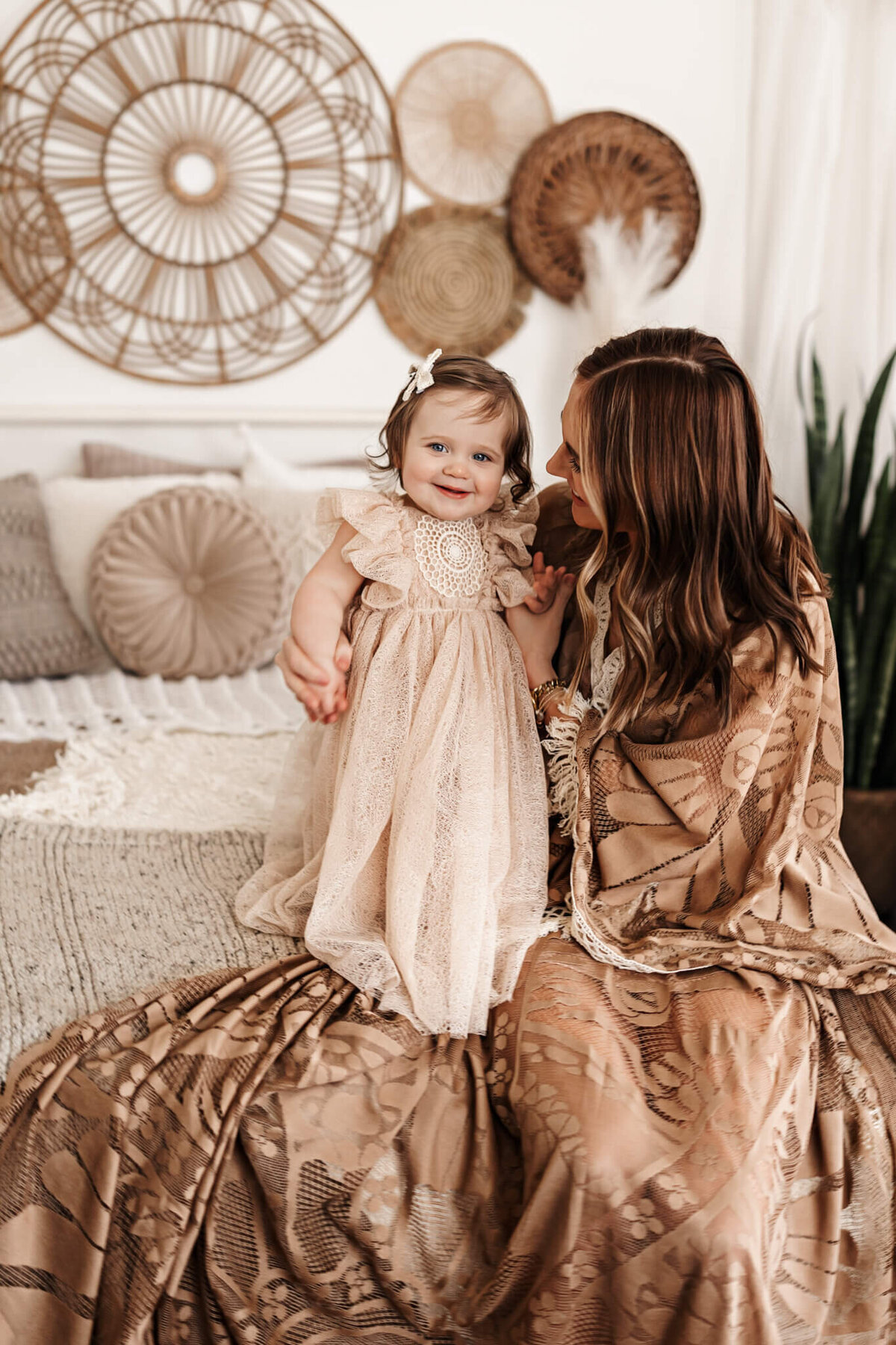 A woman and a toddler in matching beige lace outfits sit on a bed, captured beautifully by a portrait photographer in Bloomington, Illinois. The toddler stands, holding the woman's hand. Round wicker wall decorations behind them add to the cozy and neutral-toned setting of this lovely family photography moment.