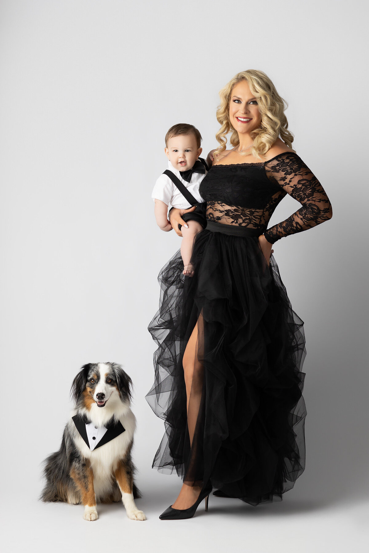 Dallas milestone photographer captures a glamorous mom in a black lace gown holding her baby dressed in suspenders and bow tie, with their dog in a tuxedo sitting at her feet.