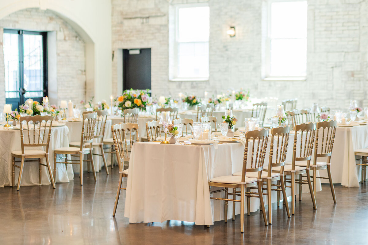 A bright wedding reception room with white brick walls, round tables covered in white linens and pastel floral centerpieces