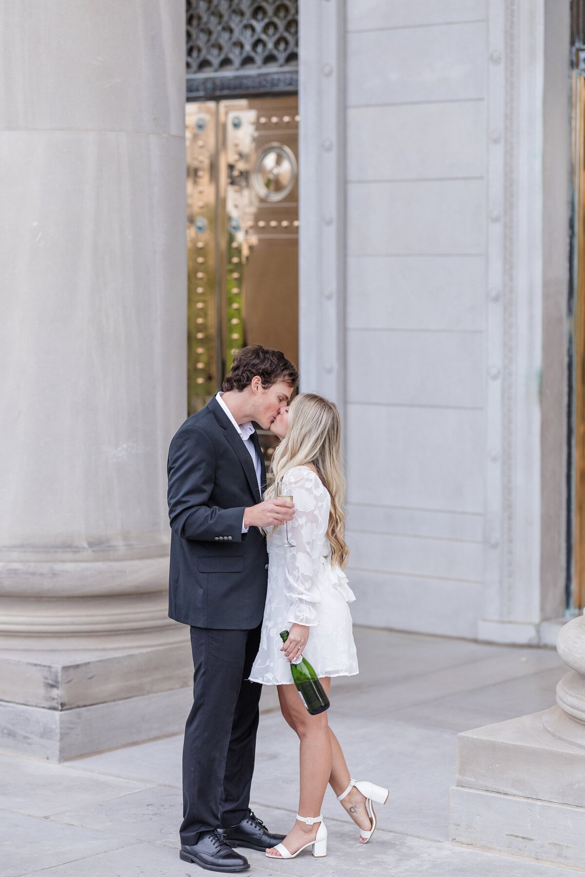 An engaged couple kisses while standing by a column as she holds a bottle of champagne
