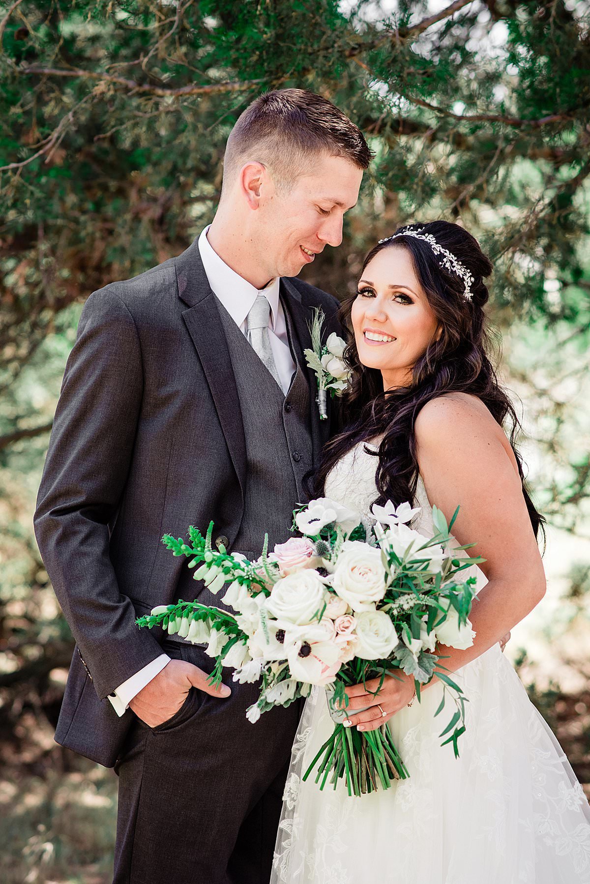 Bride holding a large bouquet with anemones, garden roses and greenery smiling at the camera and her husband is smiling at her