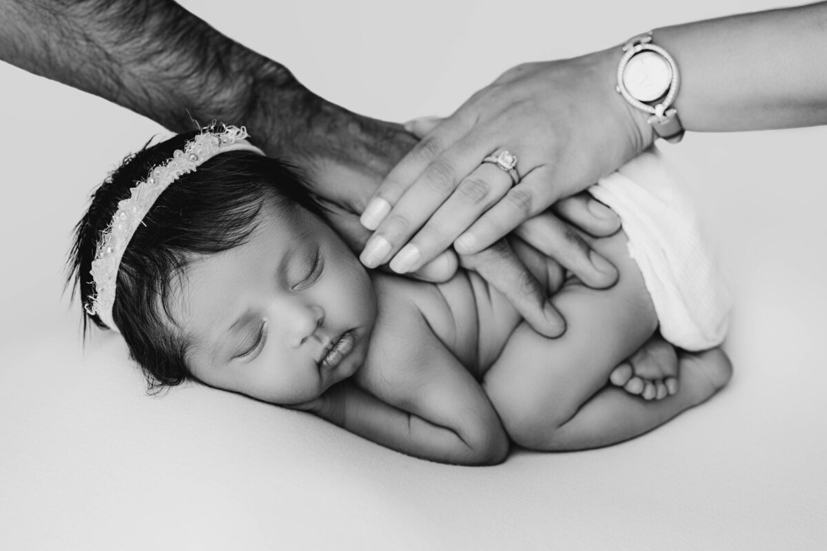 Black and white image of a newborn girl with thick hair sleeping on curled up while both parents gently rest their hands on her.