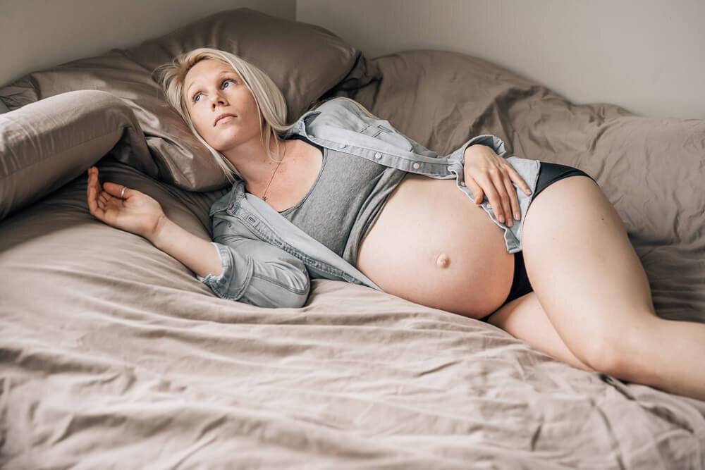 An expecting mother lounges on her bed, gazing out the window, during a bouidoir style maternity photography session.