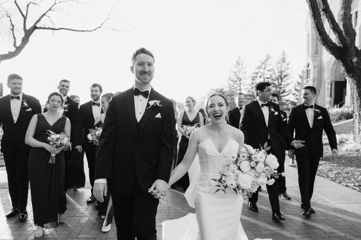 Black and white photo of a bride wearing a strapless dress with a bow and groom with a black tie tux walk hand in hand. Photo by Anna Brace, an Nebraska wedding photographer.