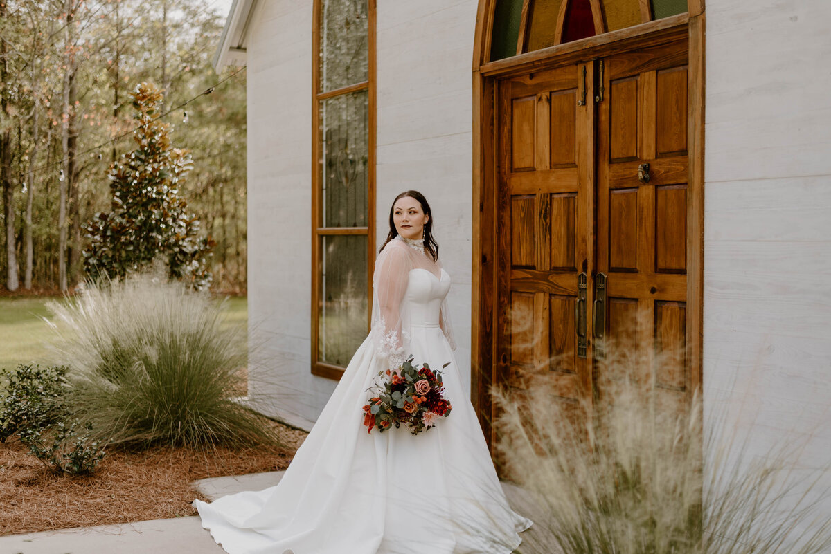 a bride in wedding dress holding her bouquet standing next to a stained glass door at cypress grove open air chapel in louisiana.