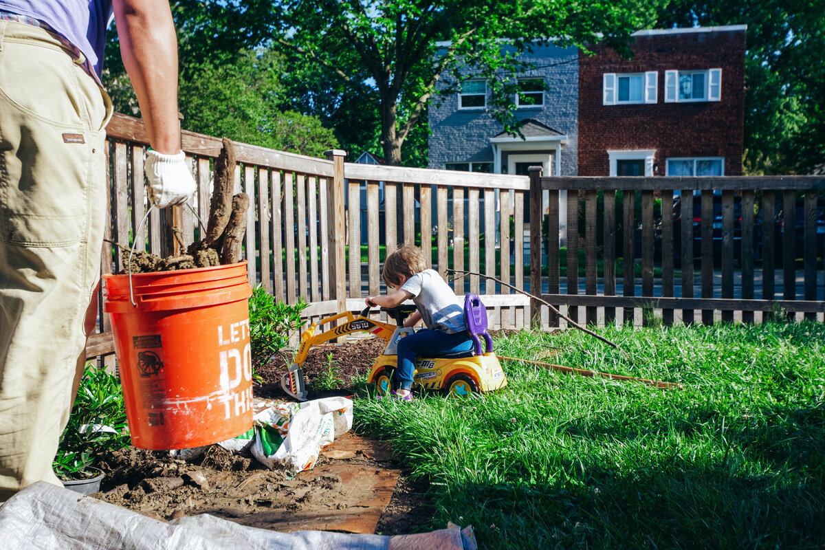 a boy digs in the yard while his dad carries a bucket of yard waste