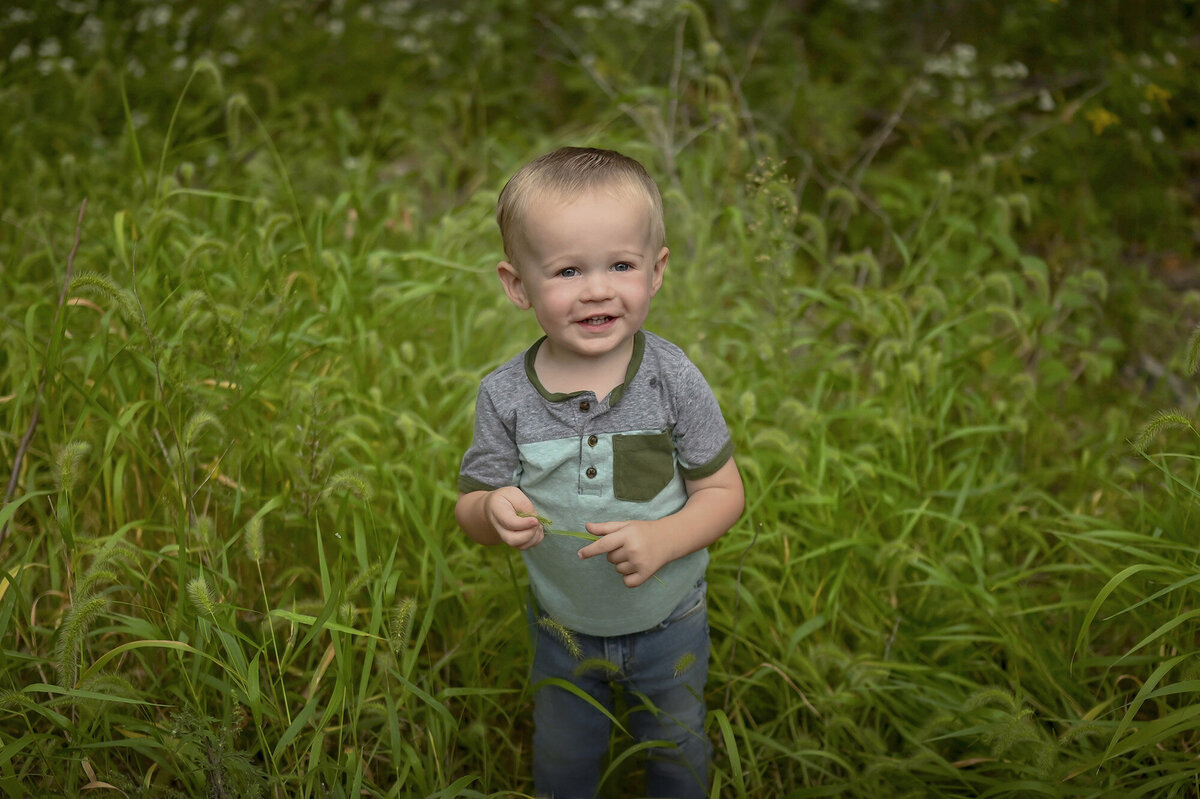 A little boy with a blue and grey shirt on holds a blade of grass