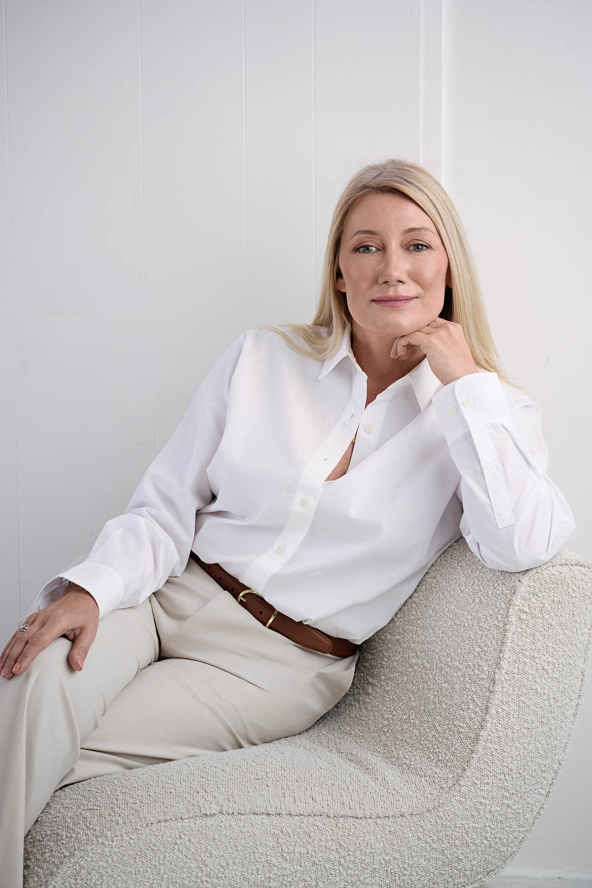 Woman in white shirt and cream pants on cream boucle couch. She's blonde with her hand against her chin looking at camera.
