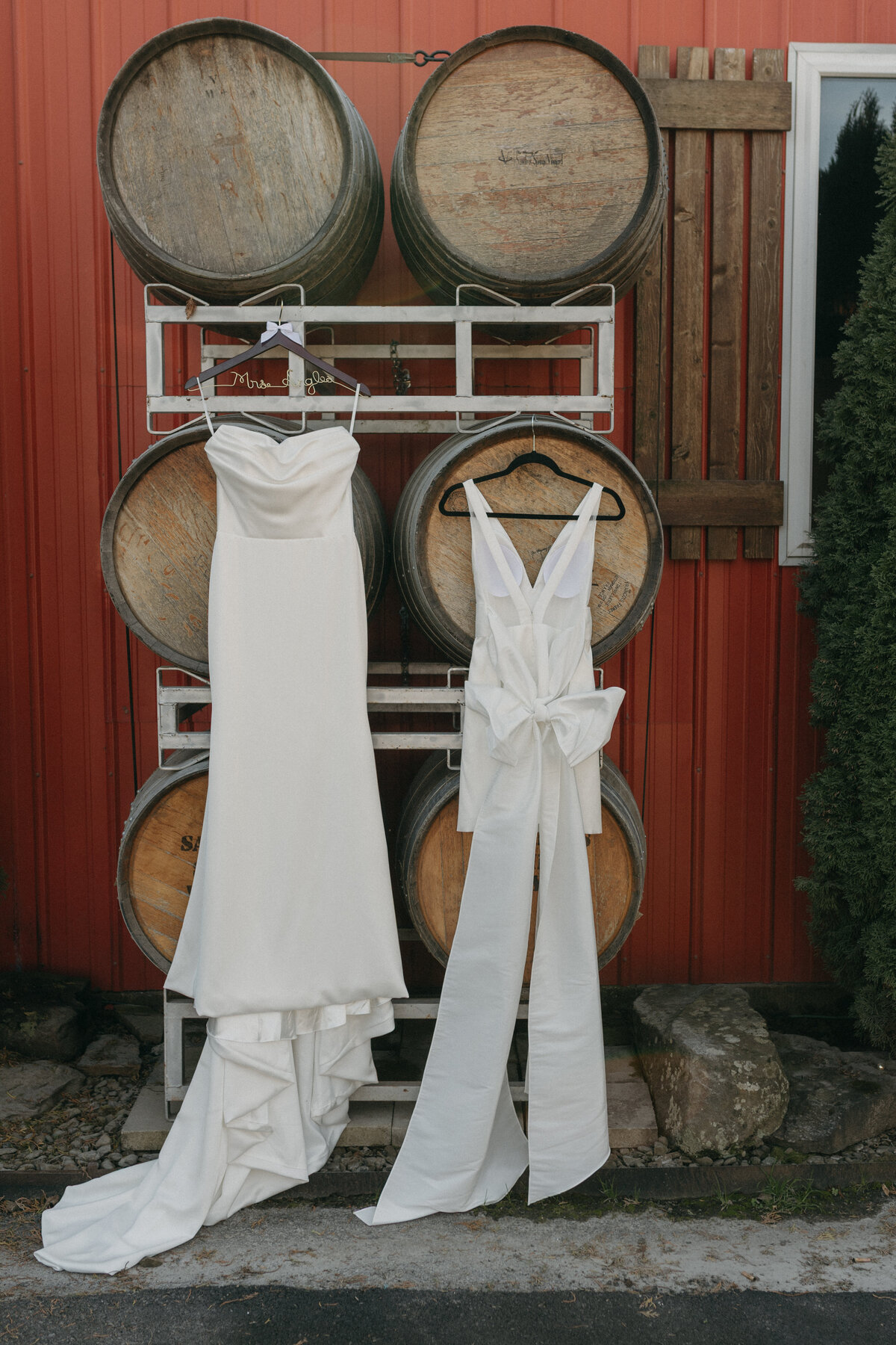 Elegant bridal gown and modern white reception dress hanging against rustic wine barrels at an outdoor wedding venue, offering inspiration for winery wedding styling and bridal fashion details.

