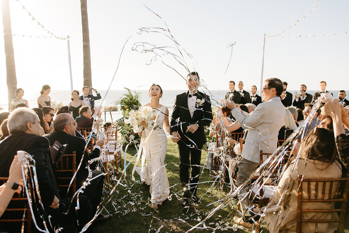 Kohnur Joyful recessional with bride in natural radiant makeup and a sleek low bun wearing a modern gown, walking down a coastal lawn with confetti.