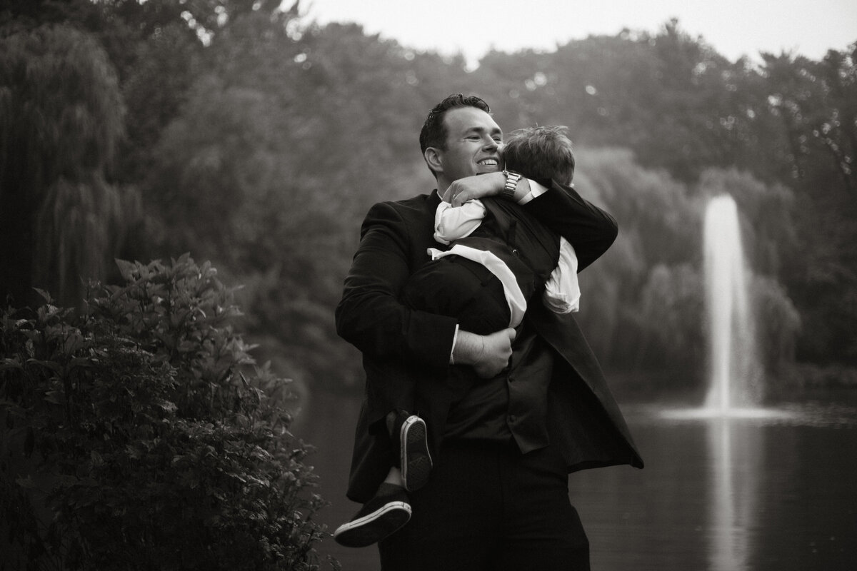 Emotive black and white photo of a father hugging his son near a fountain at a Michigan City, Indiana wedding, captured by Rainstorm Photo & Video in a heartfelt, documentary style.
