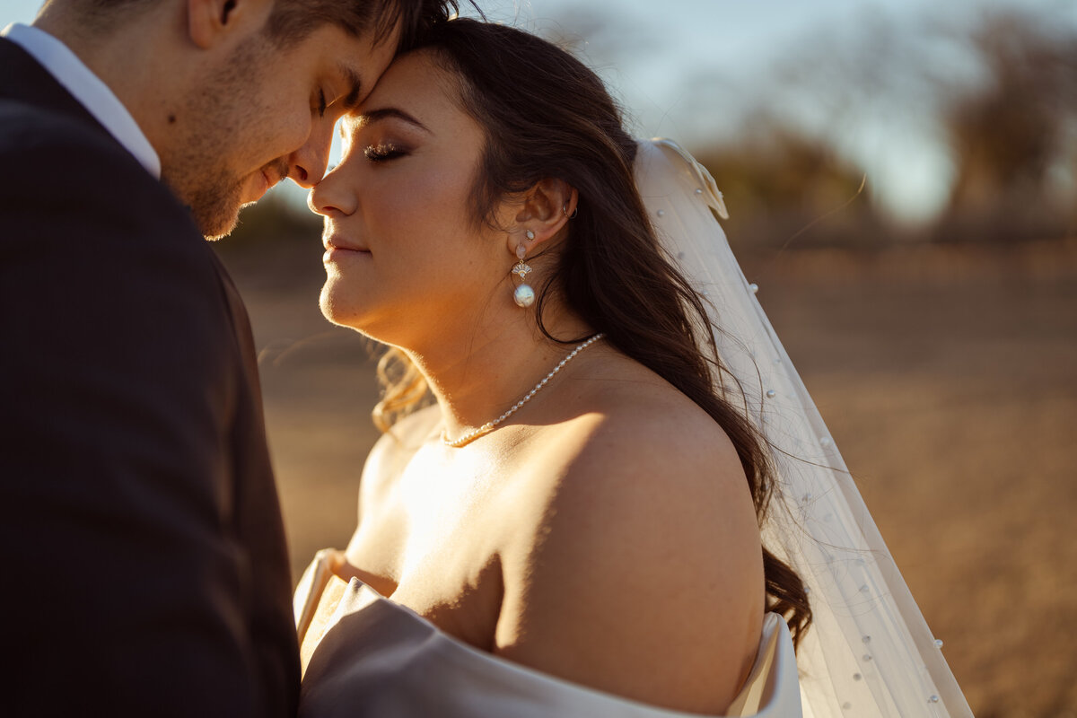 Close-up of a bride and groom sharing a close moment, with their foreheads gently touching during a winter wedding at D'Vine Grace Vineyard in McKinney, Texas. The warm sunset light highlights the bride's peaceful expression and elegant pearl jewelry.