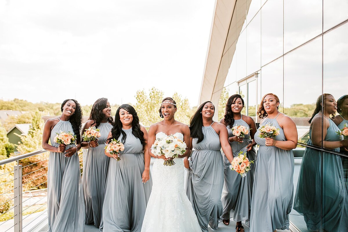 An African American bride wearing a strapless wedding dress and rhinestone headband holds a white floral bouquet with light orange roses. Standing on either side of the bride are wearing silver gray halter top satin bridesmaid dresses. The bridesmaids are holding bouquets of light orange and white roses at The Liff Center in Nashville.