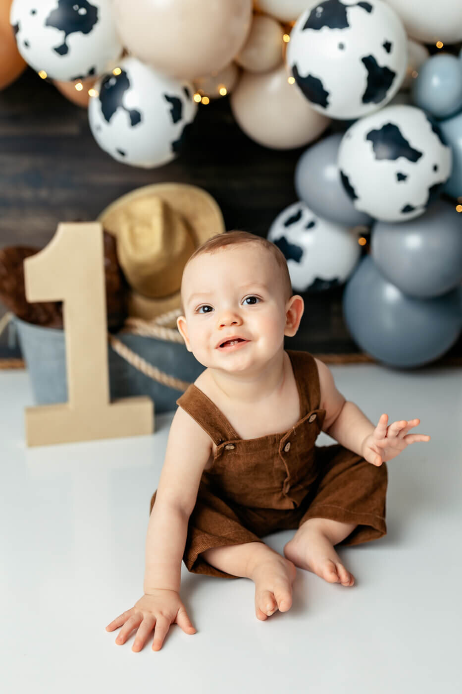 A baby wearing brown overalls sits on the floor, smiling. Behind is the number one, a cowboy hat, and a cluster of black, white, and beige balloons with cow patterns. Small lights are intertwined with the decorations.