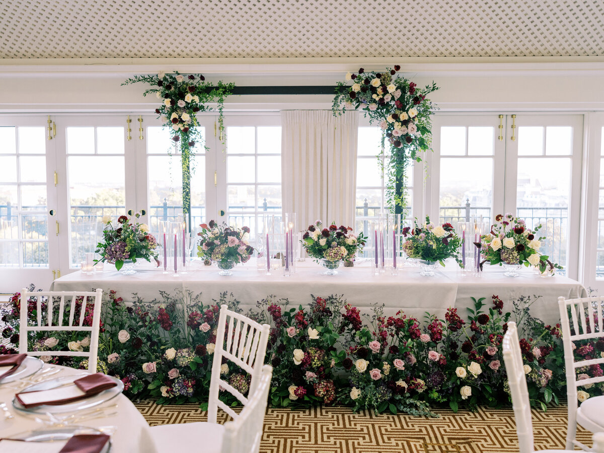 head table with jewel toned centerpieces and candles, an arch behind the table and a floral meadow in front of the table