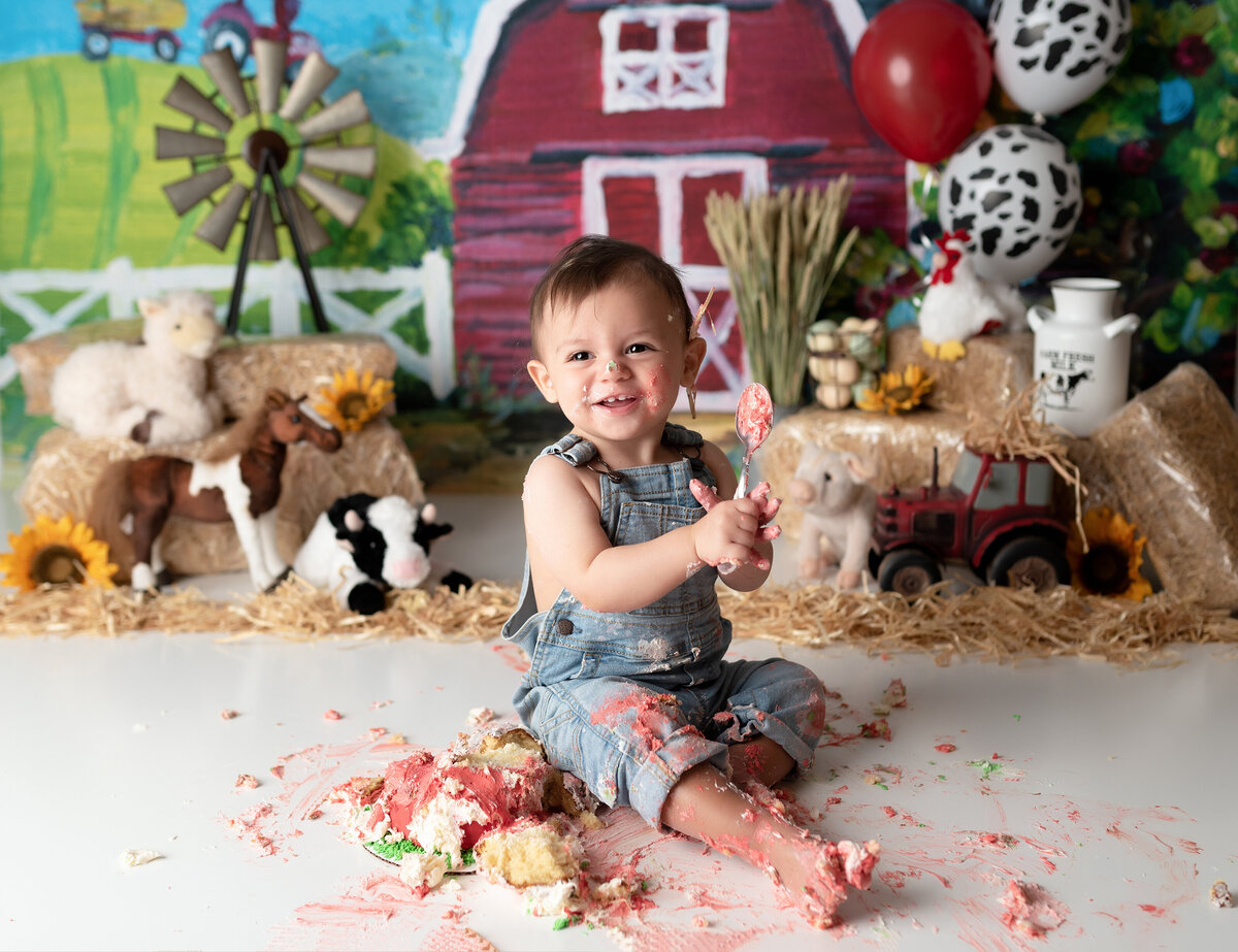 Farm themed cake smash at West Palm Beach and Delray Beach, FL photography studio. Baby boy is wearing overalls and cake is smashed on the ground  and covers his overalls. Baby is smiling at the camera. In the background, there is a farm backdrop, hay, vintage tractor toy, and stuffed farm animals.