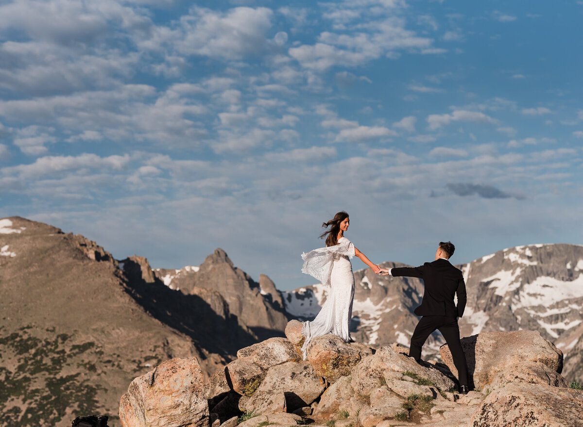 A photo of a bride and groom reaching for each others hands with large mountains in the background