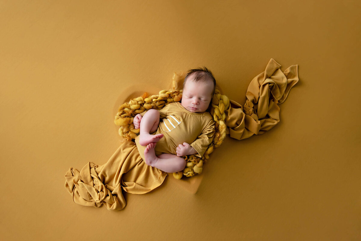 newborn boy wearing a yellow onsie in a heart-shaped bown on a yellow background for his newborn photography session.