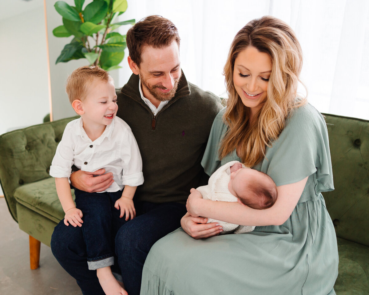 Family with their newborn on a dark green sofa. The mother, in a long light green dress, holds the baby while the man and older boy smile at the baby