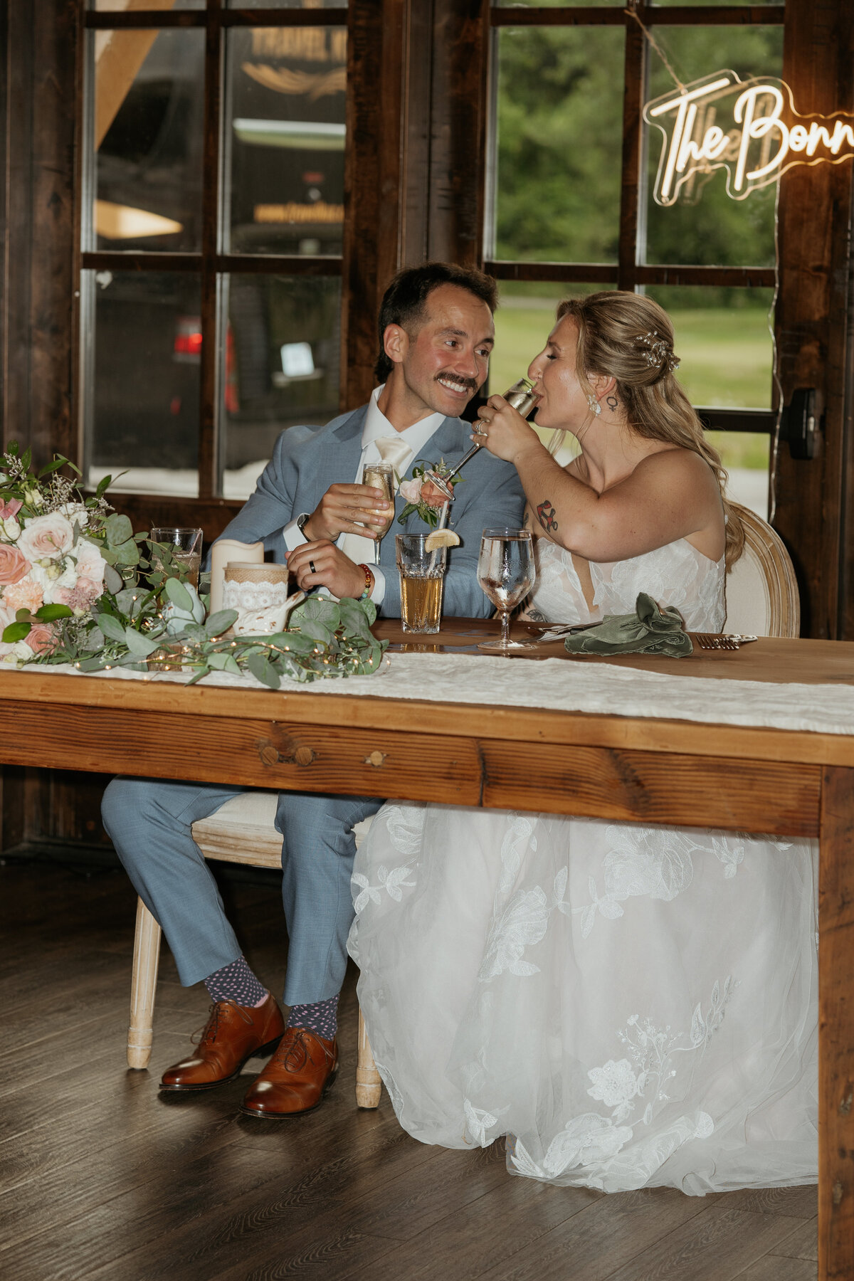 Bride and groom share a sweet moment during their wedding reception, toasting with drinks while seated at a beautifully decorated table. The bride, in a lace gown, and the groom, in a light blue suit, smile at each other under a neon sign that reads 'The Bonnys,' capturing the joy and love of their celebration.