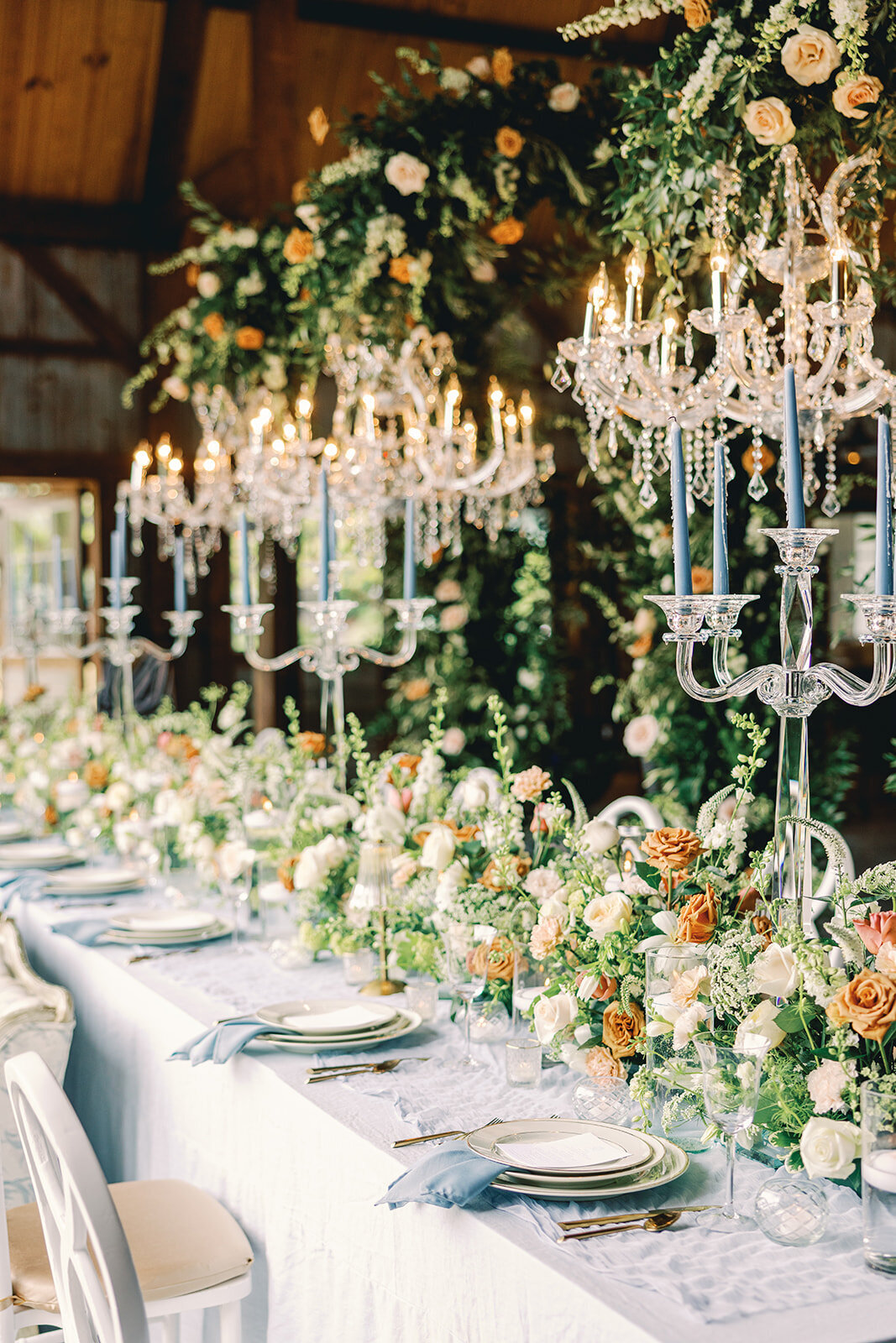 Elaborate table setting with large orange and green floral centerpieces, blue candles and floral arrangements hanging in the chandeliers