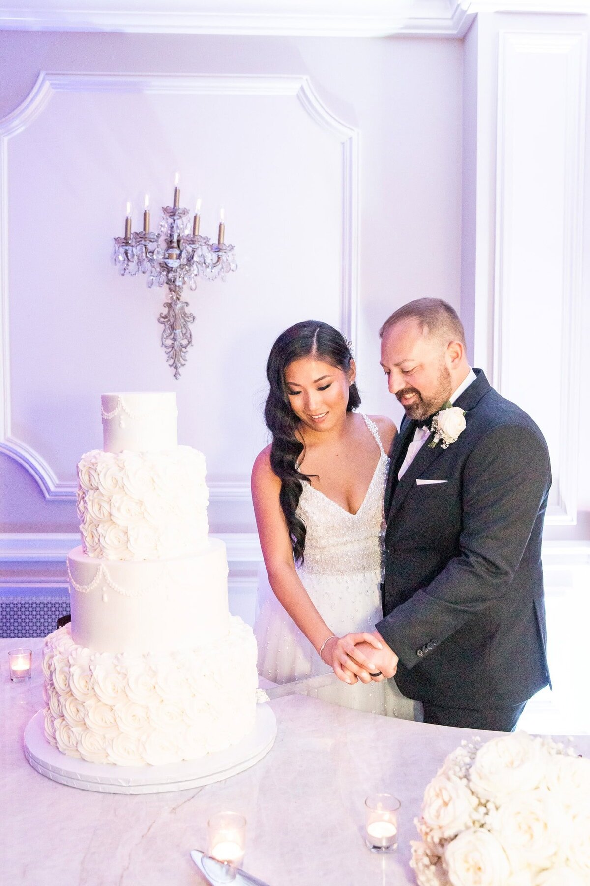 The newlyweds stand side by side, hands intertwined as they slice into a towering white wedding cake adorned with intricate rosettes and pearl-like accents. Soft candlelight flickers across the table, illuminating the bride’s beaded gown and the groom’s classic black tuxedo with a crisp white boutonniere