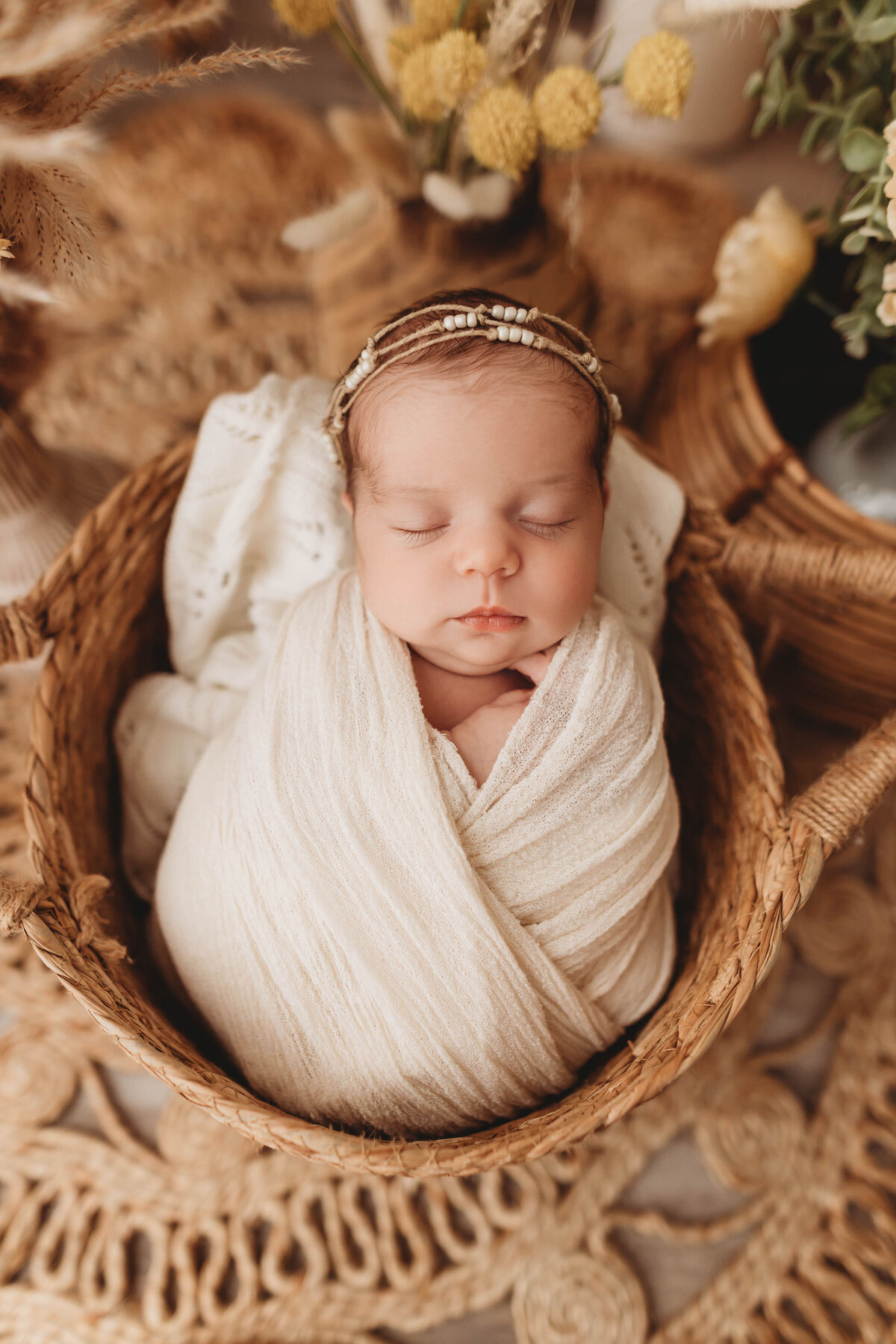Newborn baby girl wrapped in a cream blanket wearing a dainty headband in a wooden basket.