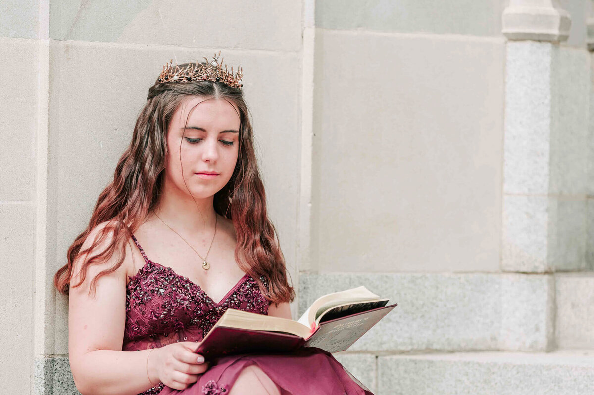 Sitting on stone steps looking at a book of fairy tales and wearing a tiara. 