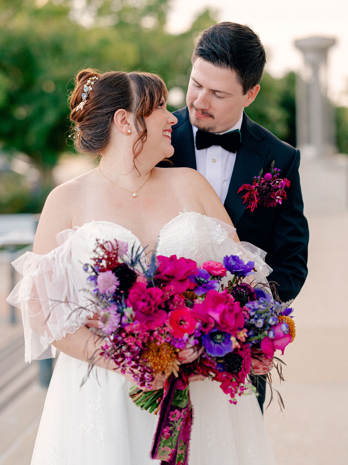 A groom hugs his bride from behind. They are looking at each other, and she is holding a large bouquet of jewel-toned flowers.