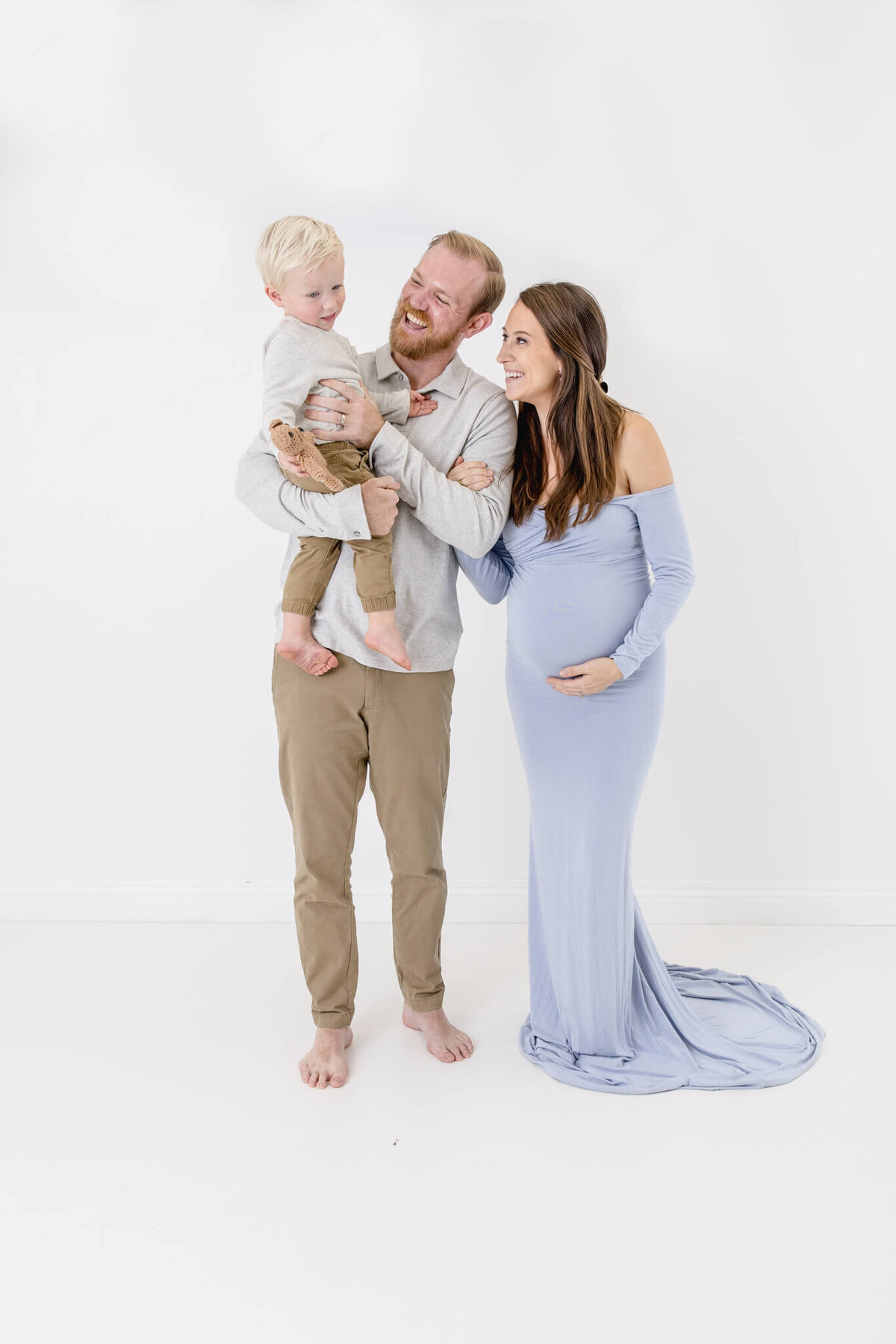 A smiling family standing against a white background. A man holds a child in his arms, and a pregnant woman in a long, light blue dress stands beside them, looking at each other fondly.