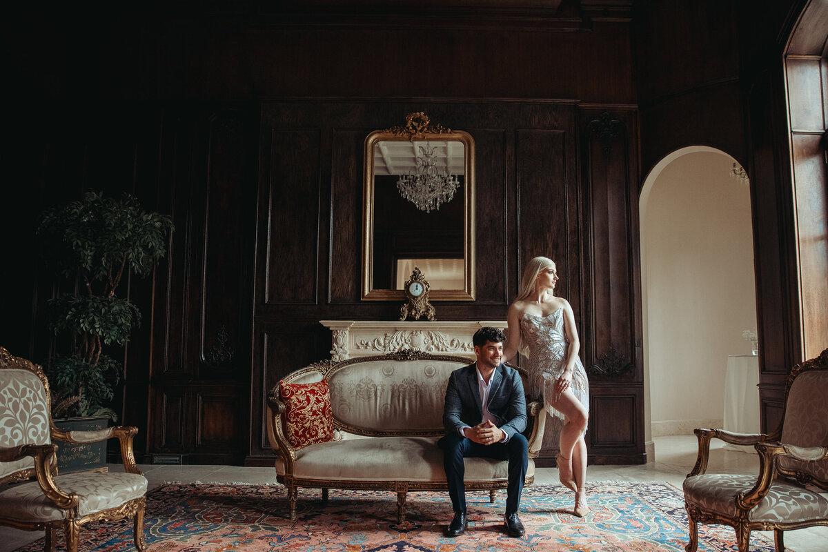 Couple in formal attire posed on a vintage sofa in a moody, wood-paneled room with chandelier.