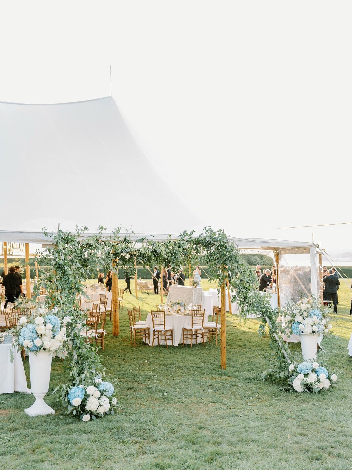 Newport Wedding Photographer | Outdoor wedding reception with a white canopy tent, surrounded by greenery and floral arrangements. Wooden chairs are arranged around tables