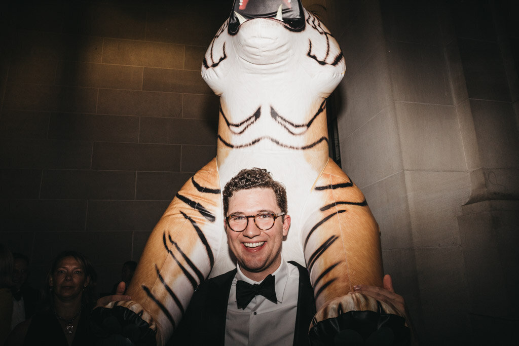 This is where the photo was taken at Washington D.C., The Mellon Auditorium, one of the best luxury DC venues with European flair, captured by photographer Sarah Bradshaw. In the scene, a man with short curly hair, glasses, and a black tuxedo is smiling and standing in front of a large inflatable tiger. The indoor setting features low lighting and stone walls in the background.