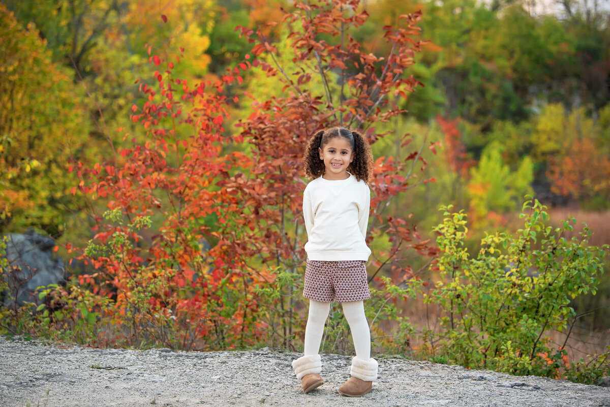little girl standing on a cliff in front of fall foliage for her family photography session in Burlington