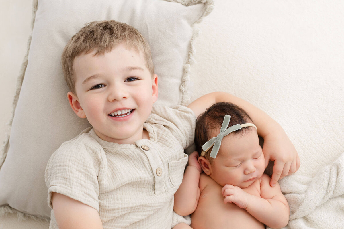 Toddler boy laying next to new baby sister with arm around her. Brother is smiling up at the camera, baby is sleeping.