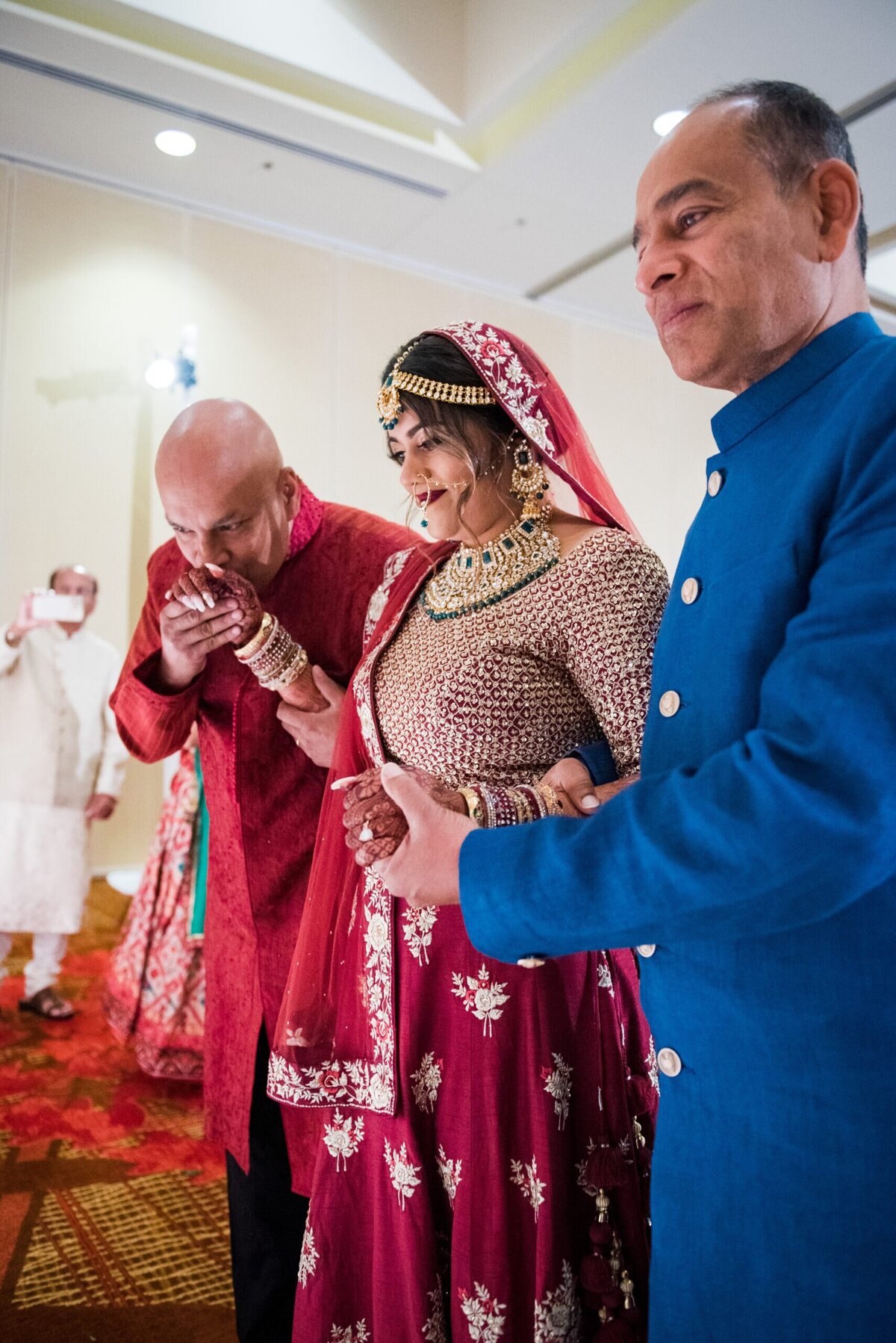 An Indian bride and her two father figures stand with her at the end of the aisle. One of the men kisses her on the hand.