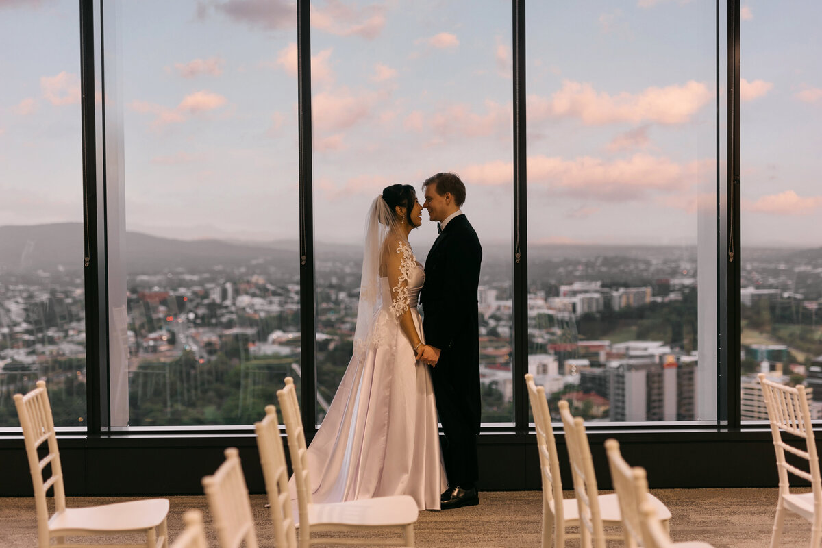 Bride and groom standing inside the Brisbane Registry Office, surrounded by ceremony chairs, with golden hour light and city views behind them.